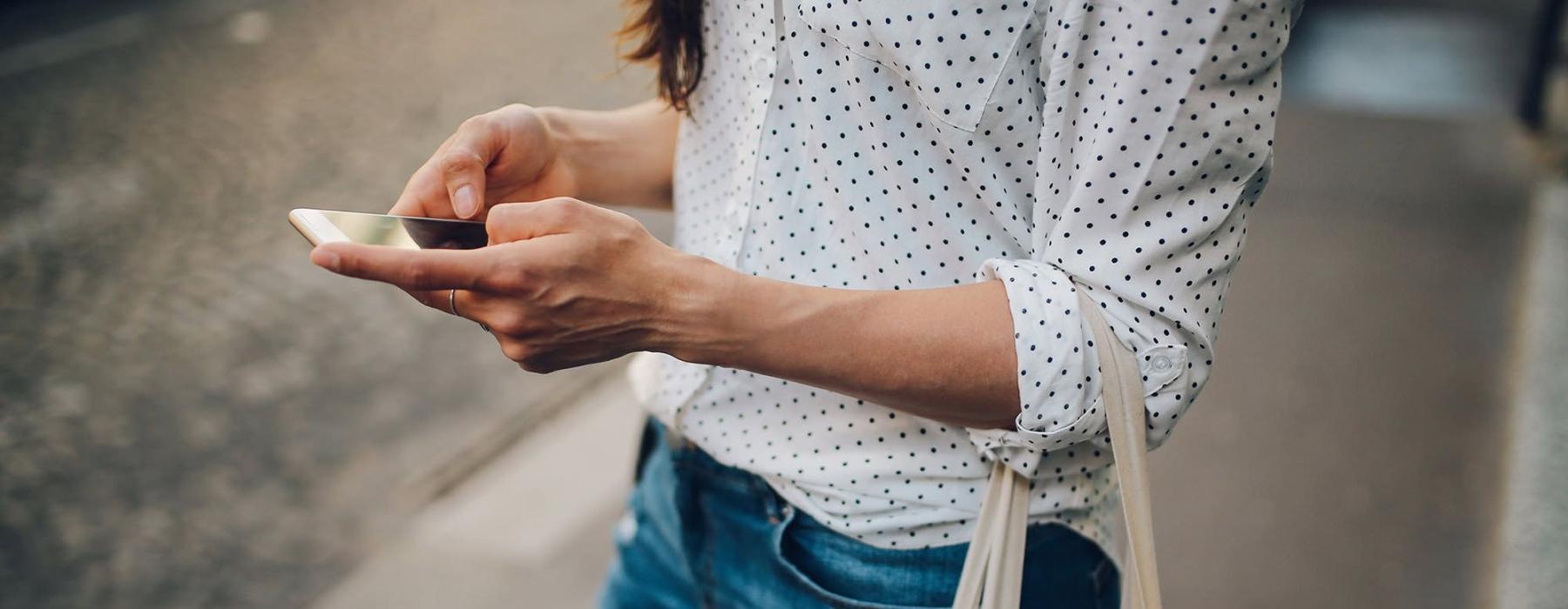 woman walks down the street and texts with a bag of groceries on her arm