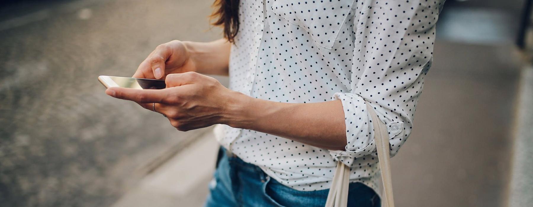 woman walks down the street and texts with a bag of groceries on her arm